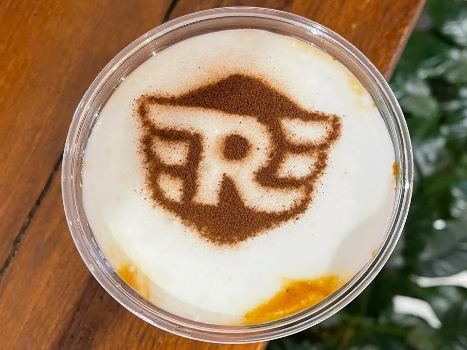 白い泡の上にココアパウダーで翼のある「R」のロゴが描かれたラテアートドリンク